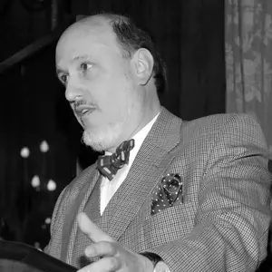 A balding man, John Simpson, with a gray beard and mustache, wearing a gray houndstooth suit, striped bow tie, and polka dot pocket square, speaks while gesturing from a lectern.