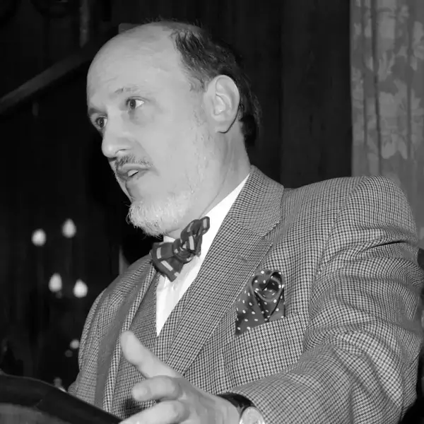 A balding man, John Simpson, with a gray beard and mustache, wearing a gray houndstooth suit, striped bow tie, and polka dot pocket square, speaks while gesturing from a lectern.