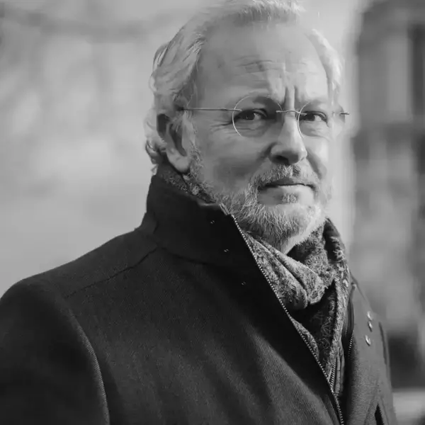 Black and white portrait of an older man, Philippe Villeneuve, with grey hair, a beard, and glasses, wearing a dark coat and patterned scarf, looking at the viewer.